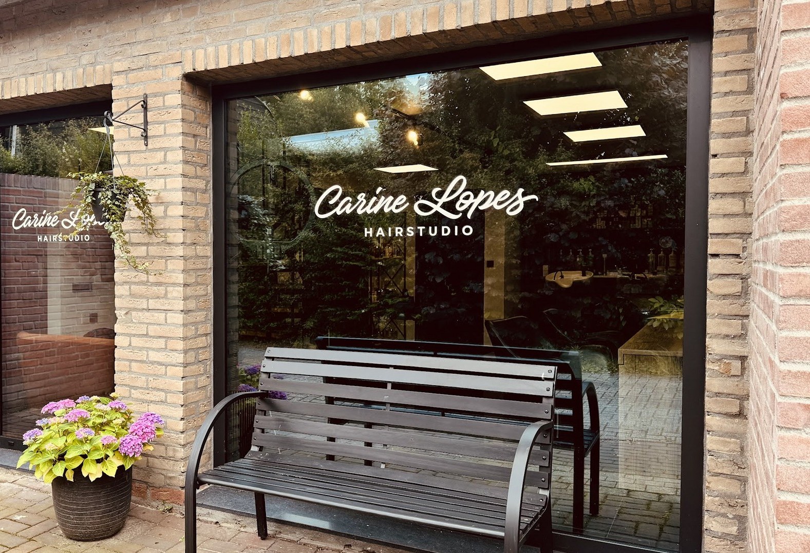 De entree van Carine Lopes Hairstudio, met het logo op het raam, een bank en hortensia’s
